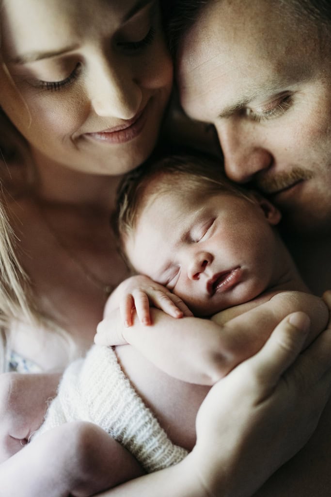 A mother and father hold their newborn baby between them. Both are gently smiling.