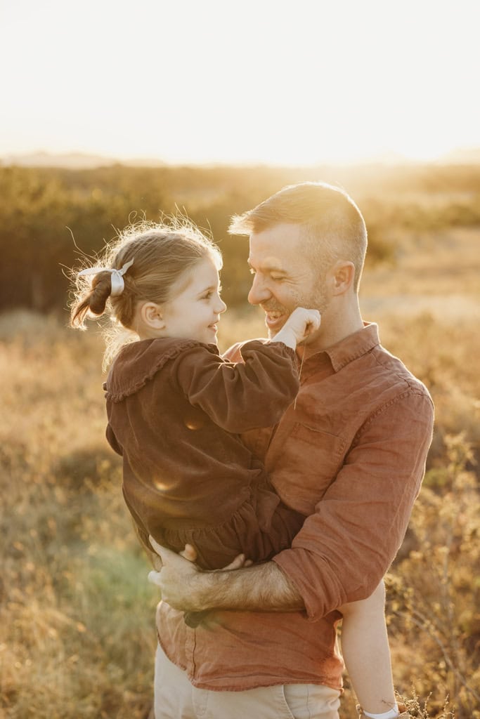 A father laughs with his daughter as he holds her and she waves a piece of grass. The sun sets behind them.
