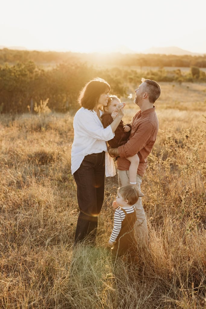 A family of four tickle each other with grass as they laugh. The sun sets behind them. 