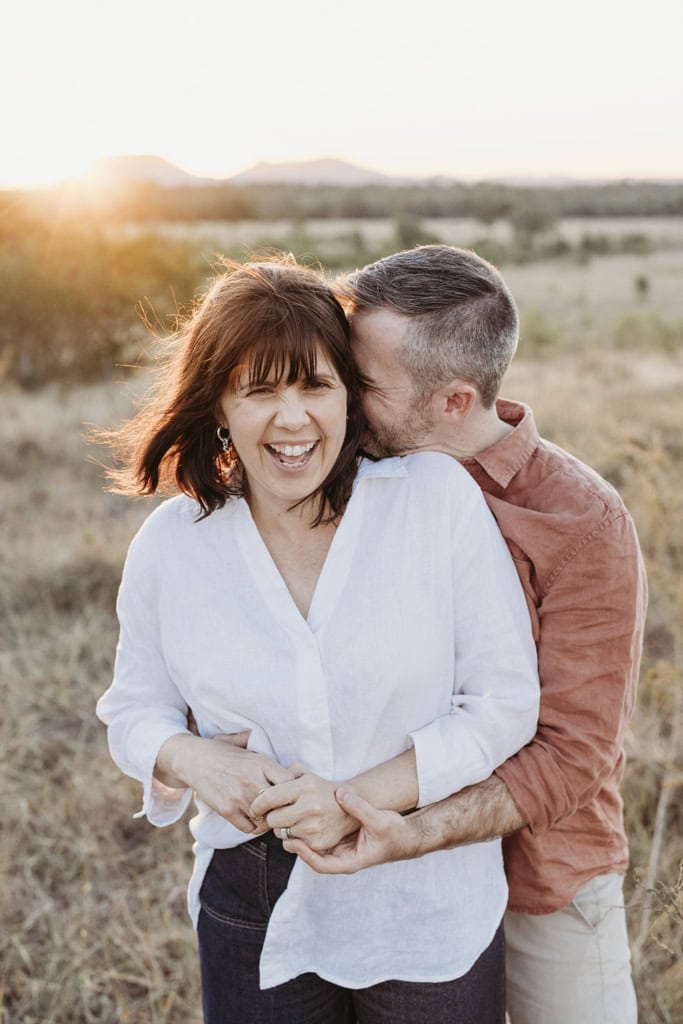 A couple laugh together as they stand in a paddock and the sun sets.