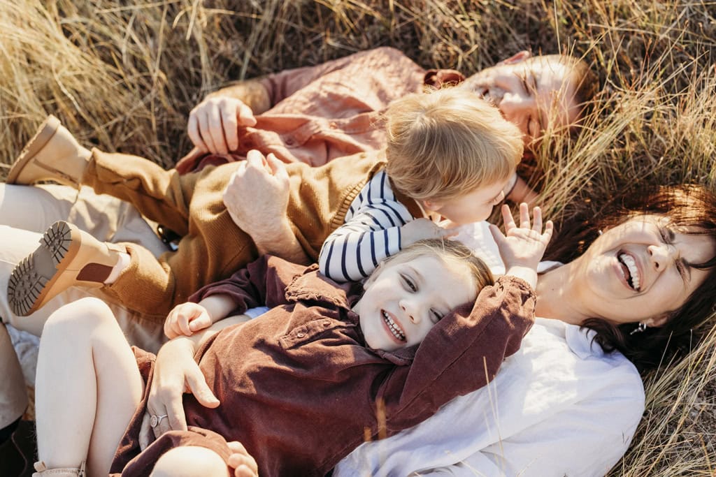 A family of four lie in long grass as they laugh and cuddle.
