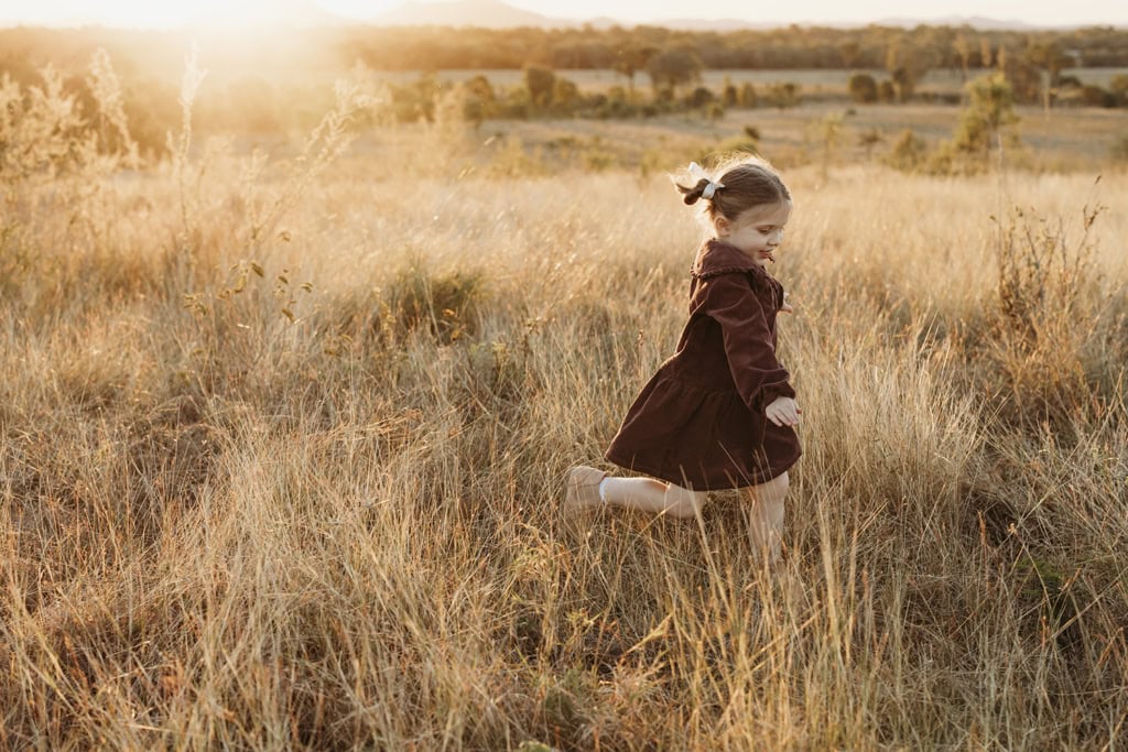 A little girl runs through long grass as the sun sets behind her.