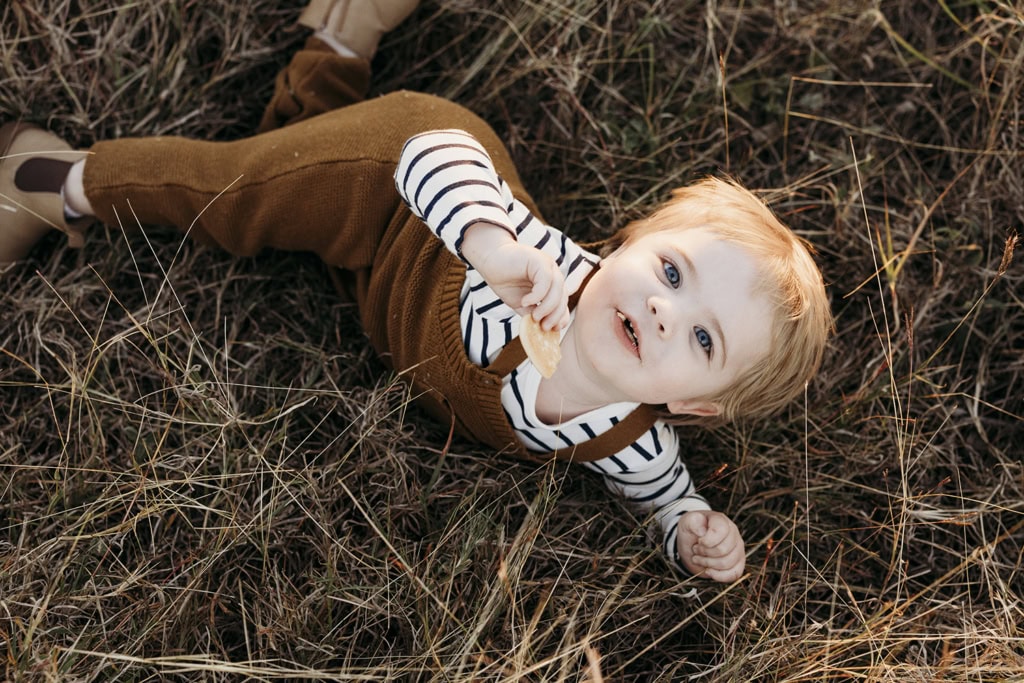 A toddler looks up at the camera as he plays in long grass.