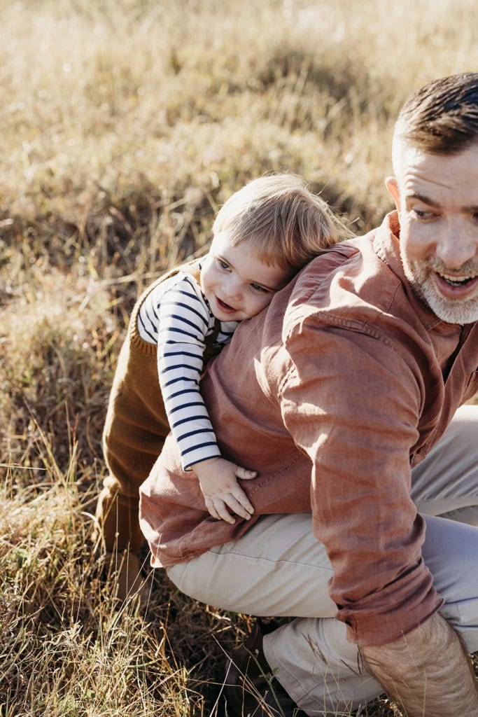 A little boy cuddles his father from behind as they both smile.
