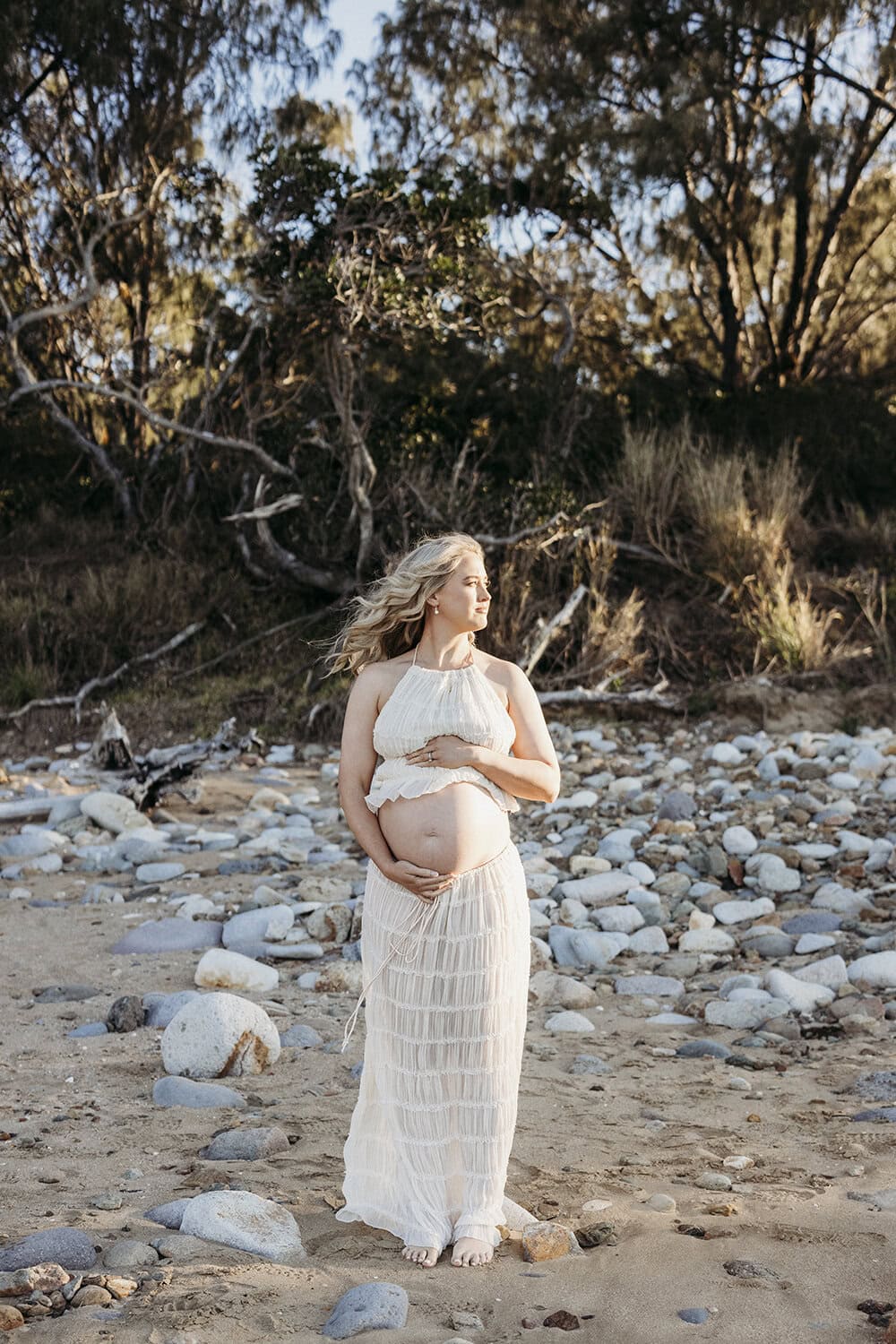 A pregnant woman stands on a beach gently cradling her belly. 