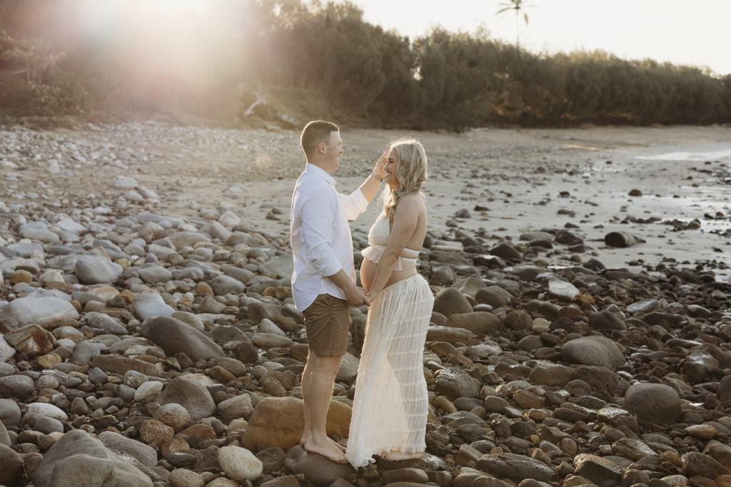 A couple face each other while holding hands, standing on a beach. The sun shines from behind them. 