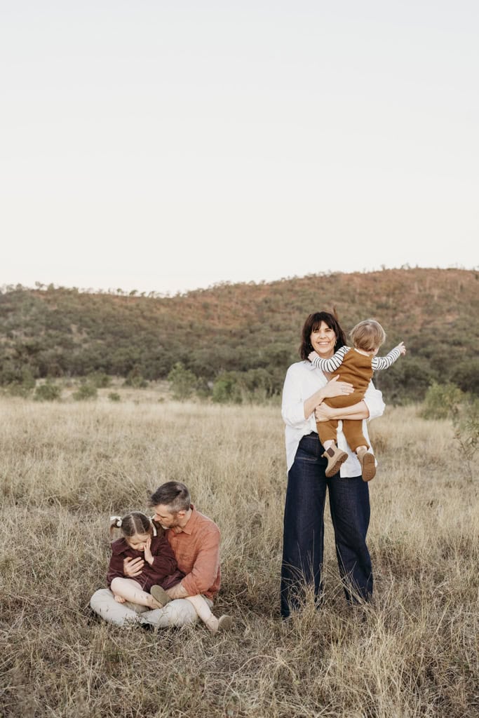 A mother plays with her toddler while her husband cuddles their daughter on the ground. They are sitting in a paddock, a mountain in the background. 