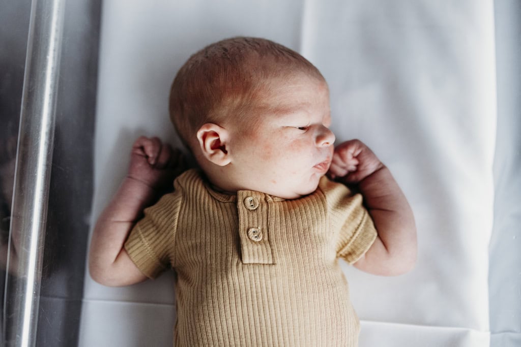 A newborn baby lies asleep with his fists clenched near his face as he lies in a clear hospital bassinet.