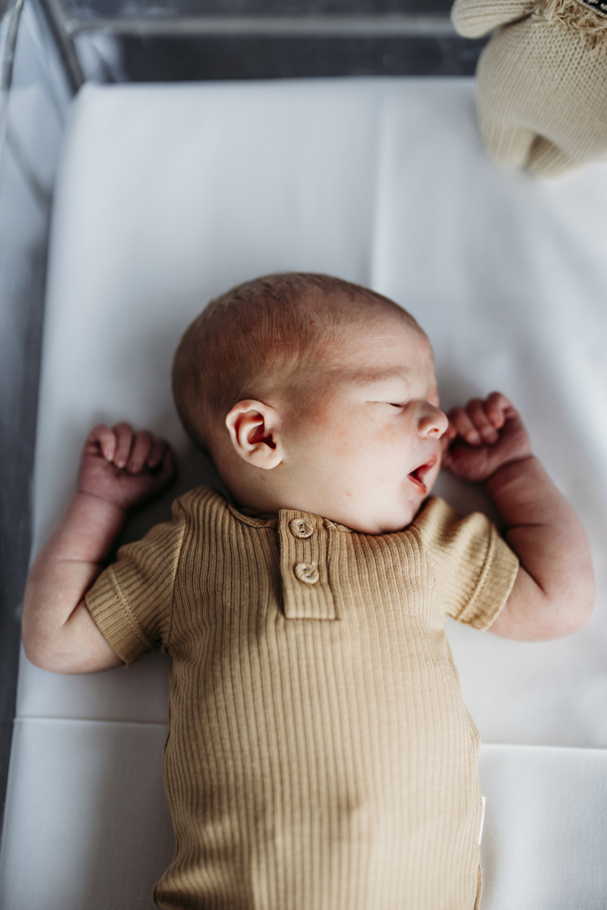 A baby lies in a clear hospital bassinet, yawning with both hands at his head.