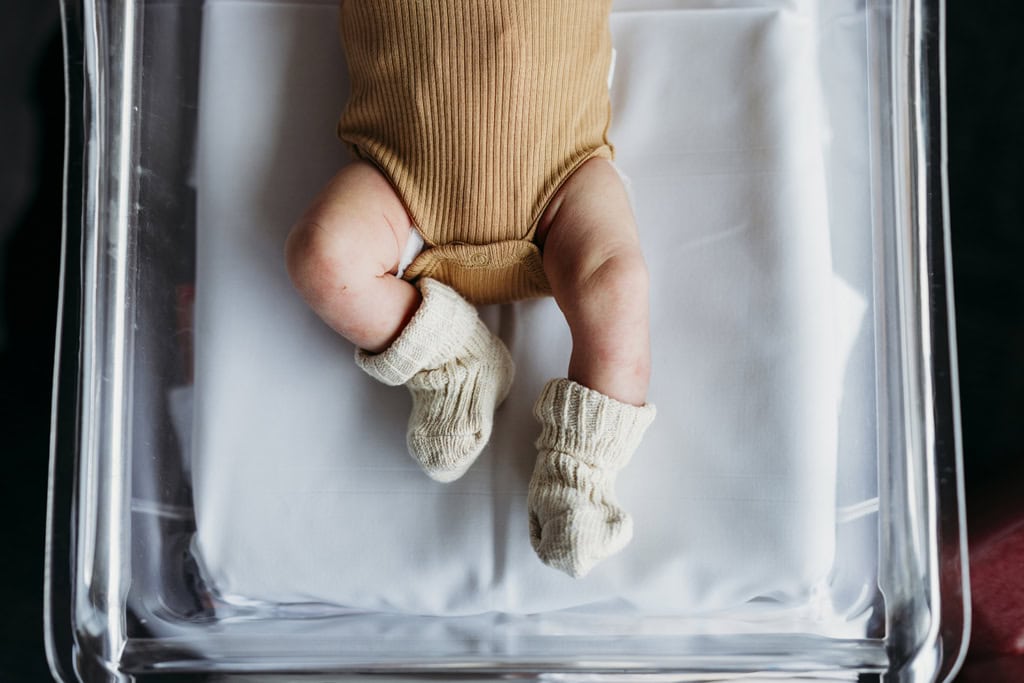 A newborn baby's legs lie in a clear bassinet in hospital. The baby is wearing cute little socks.