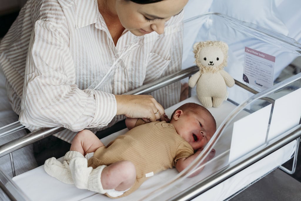 A mother leans over a clear bassinet at her newborn baby in hospital.