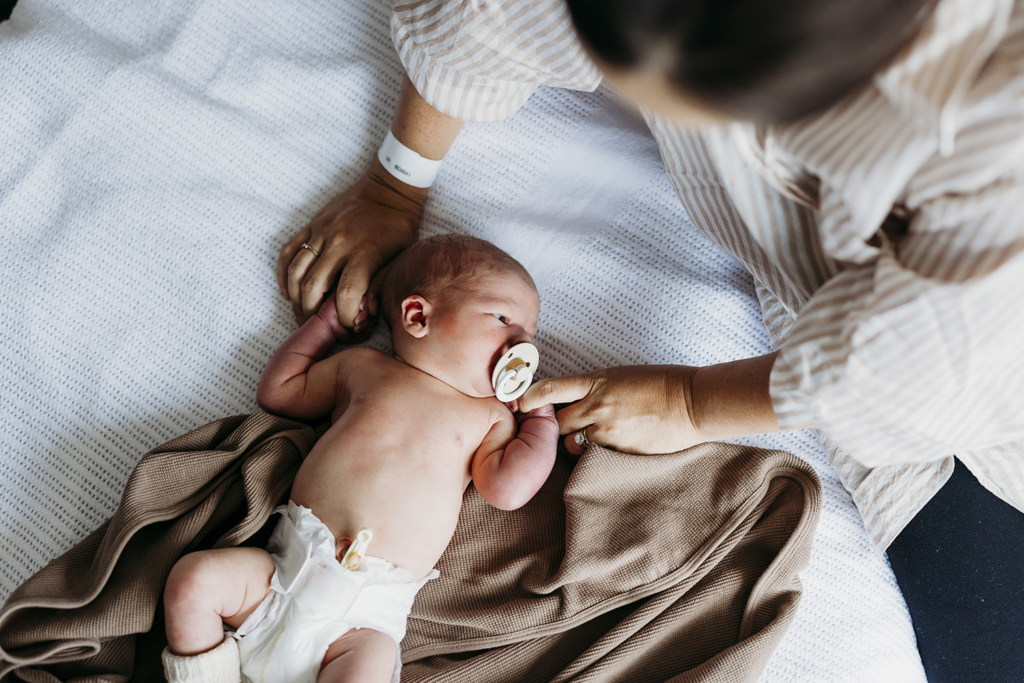 A mother holds her newborn's hands as he lies on a hospital bed, looking out the window.