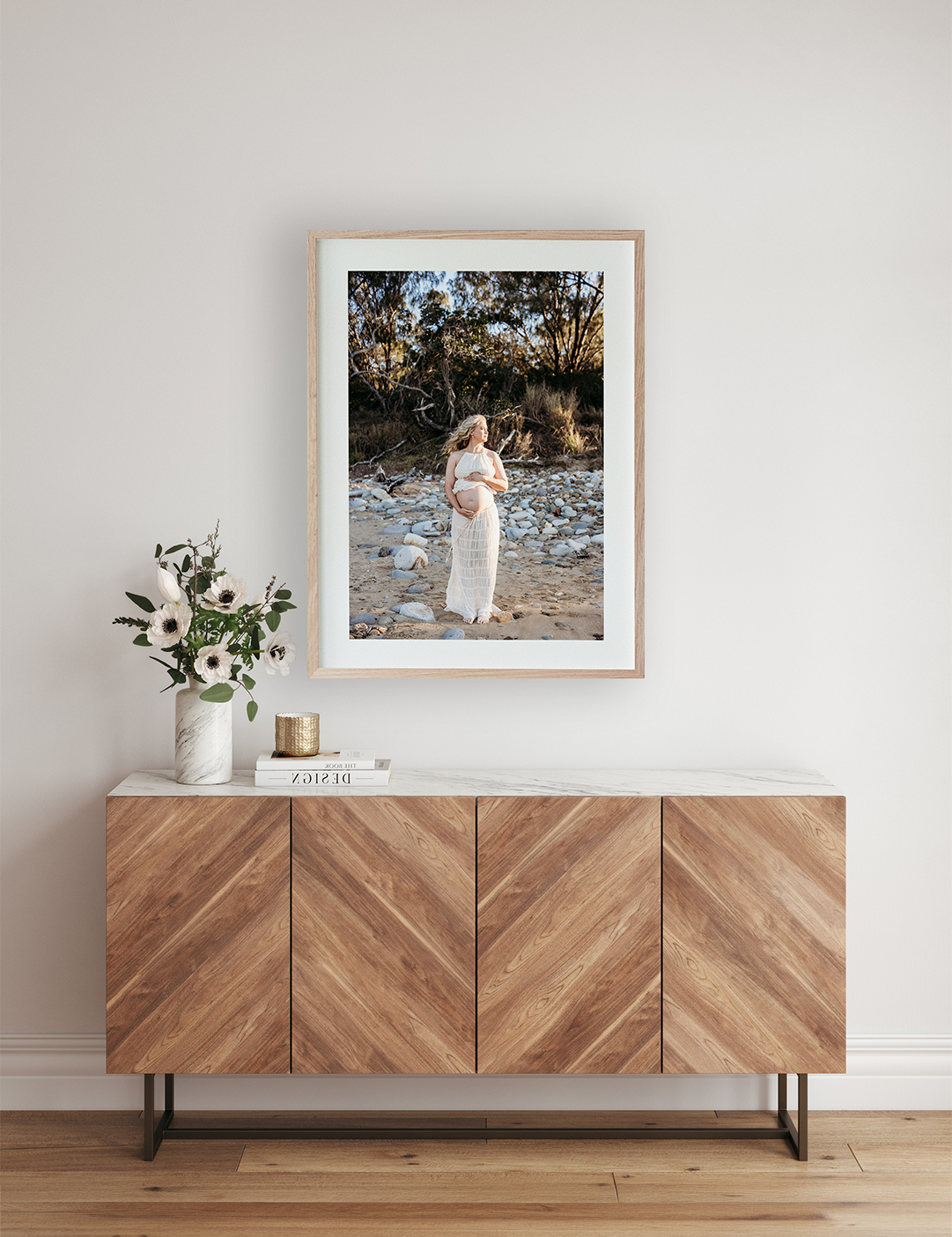 A framed portrait of a pregnant woman standing on a beach holding her belly sits above a sideboard.