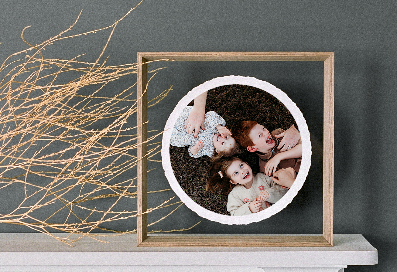 A round torn edge portrait in a clear frame with time mouldings. The photo is of three children laughing as their parents tickle them. 